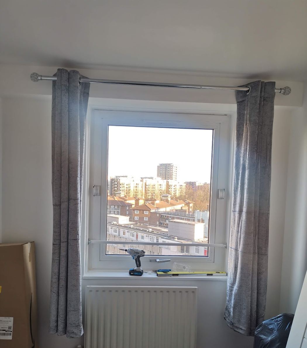 A chrome curtain pole fitted above a bedroom window with grey curtains hung, city rooftops visible through the window — installed by Swift Handy Works.
